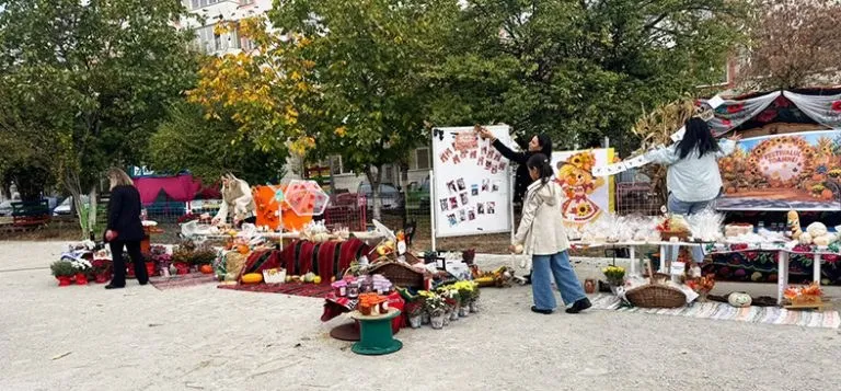 Oameni stau lângă mese cu diverse produse și decorațiuni de toamnă într-o zonă deschisă cu copaci în fundal.