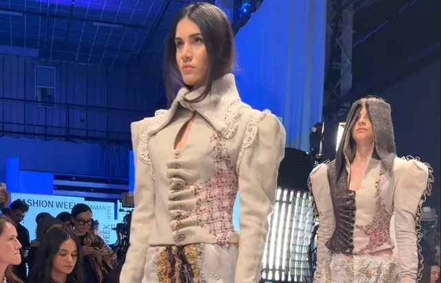 O femeie poartă o jachetă albă cu detalii decorative și broderii, mergând pe o pistă de prezentare în fața unui public.