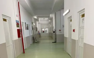 Un coridor de spital cu pereți albi, uși albe și un podea de culoare deschisă, având câteva scaune și un panou roșu pe perete.