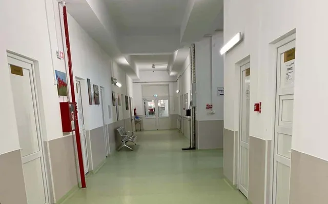 Un coridor de spital cu pereți albi, uși albe și un podea de culoare deschisă, având câteva scaune și un panou roșu pe perete.