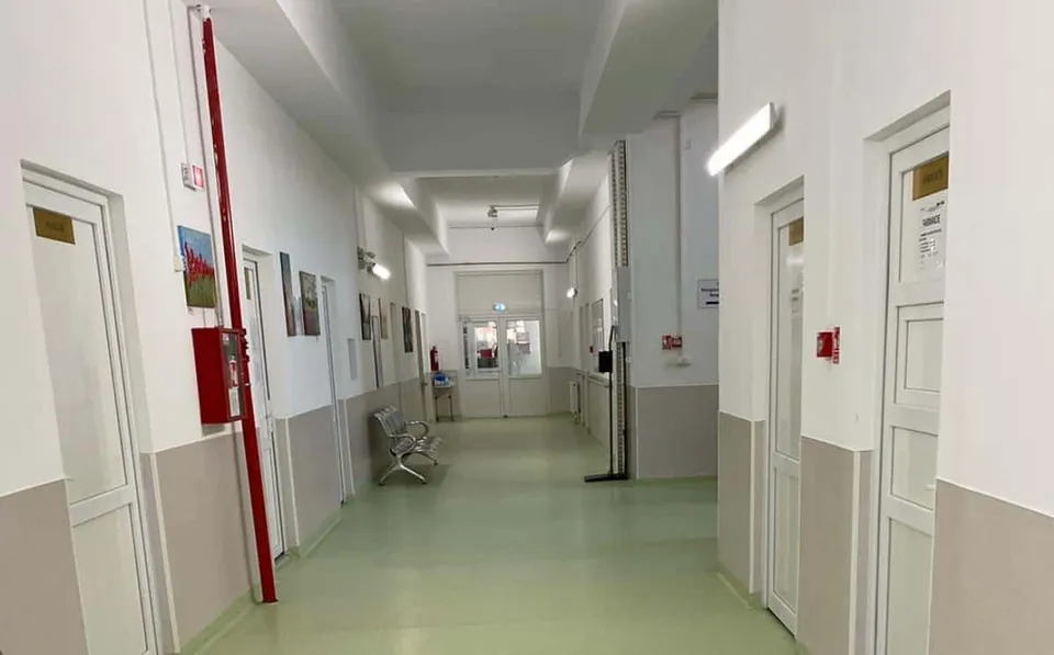 Un coridor de spital cu pereți albi, uși albe și un podea de culoare deschisă, având câteva scaune și un panou roșu pe perete.