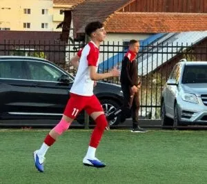 Un jucător de fotbal cu echipament alb și roșu aleargă pe un teren de iarba artificială lângă mașini și un gard metalic.