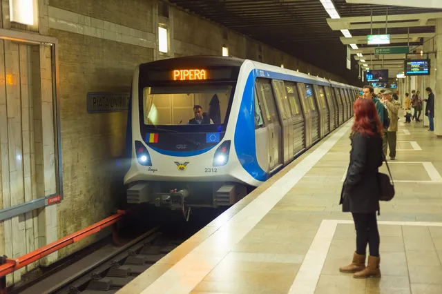 Un tren de metrou albastru și alb intră într-o stație subterană, în timp ce câțiva pasageri stau pe peronul iluminat.