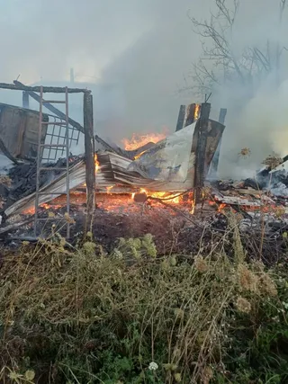 Flăcări și fum ies dintr-o structură de lemn și metal aflată în ruine, înconjuratată de vegetație uscată.