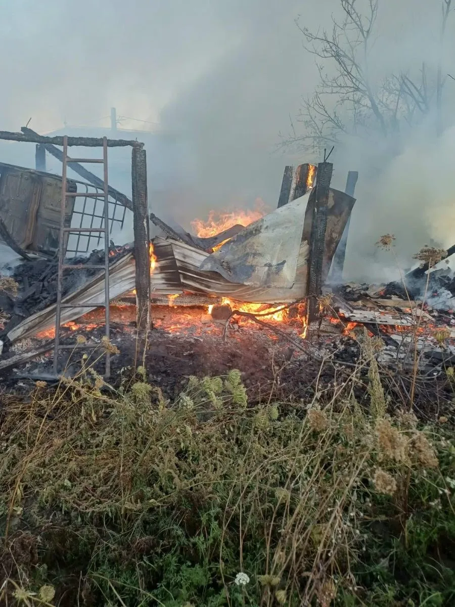 Flăcări și fum ies dintr-o structură de lemn și metal aflată în ruine, înconjuratată de vegetație uscată.