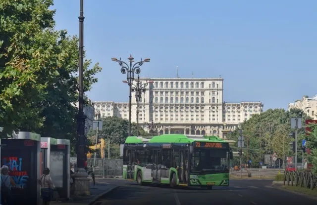 Un autobuz verde circulă pe un drum asfaltat în fața unui clădire albă masivă sub un cer senin.