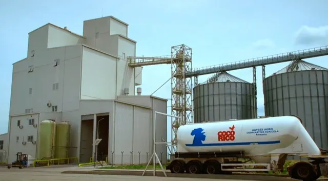 O clădire industrială înaltă, gri, este lângă două silozuri metalice mari și un camion cu o cisternă albă marcată cu diverse logo-uri.