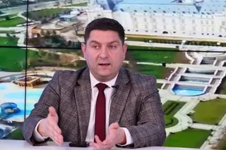Un bărbat cu păr scurt, purtând sacou gri și cravată roșie, stă în fața unui ecran care afișează o imagine aeriană a unor clădiri și grădini.