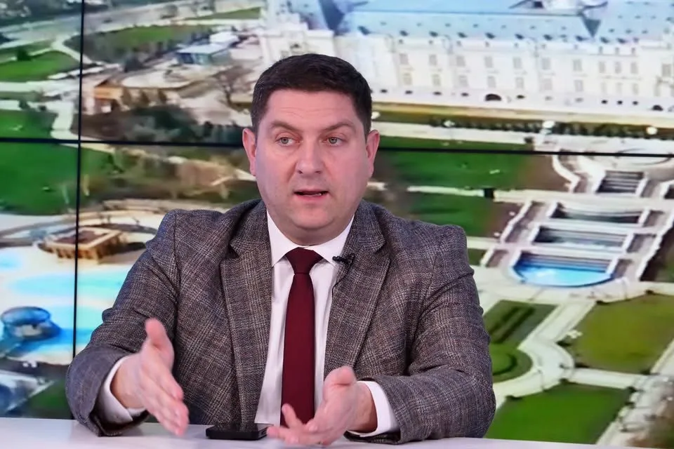 Un bărbat cu păr scurt, purtând sacou gri și cravată roșie, stă în fața unui ecran care afișează o imagine aeriană a unor clădiri și grădini.