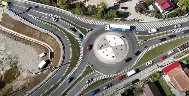 O vedere aeriană prezintă un sens giratoriu cu mai multe benzi de circulație, vehicule colorate și margini marcate cu linii albe pe asfaltul gri.