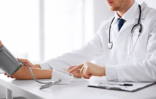 Un medic într-o halat albă folosește un tensiometru pentru a măsura tensiunea arterială pe brațul unui pacient aflat la o masă albă.
