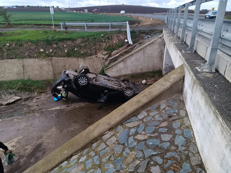 O mașină neagră este răsturnată pe oul în șanț, lângă un parapet de beton și metal de pe un pod.