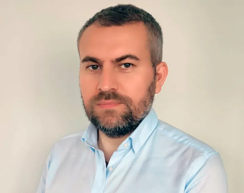 Un bărbat cu barbă și păr scurt, purtând o cămașă albastră deschisă, privește spre cameră în fața unui fundal neutru.
