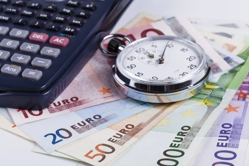 Un cronometru argintiu este așezat peste mai multe bancnote de euro și lângă un calculator de culoare închisă.