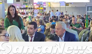 Un grup de persoane stă într-o încăpere luminoasă cu mulți oameni în fundal, printre care un bărbat cu părul cărunt și o femeie cu părul lung și negru.