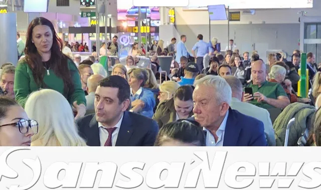 Un grup de persoane stă într-o încăpere luminoasă cu mulți oameni în fundal, printre care un bărbat cu părul cărunt și o femeie cu părul lung și negru.