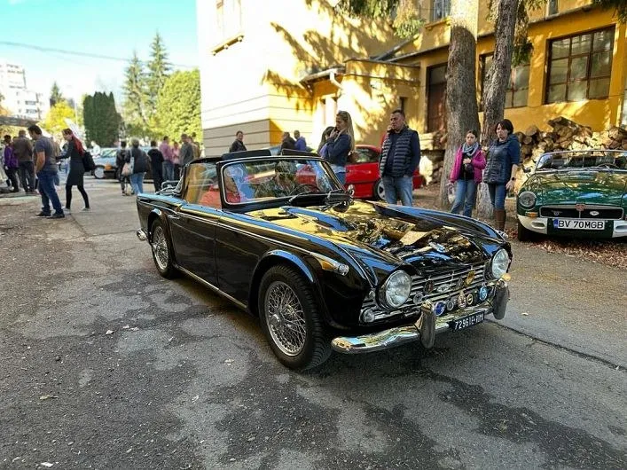 Un autoturism negru cu capotă deschisă este parcat pe un drum asfaltat lângă un grup de oameni și clădiri.