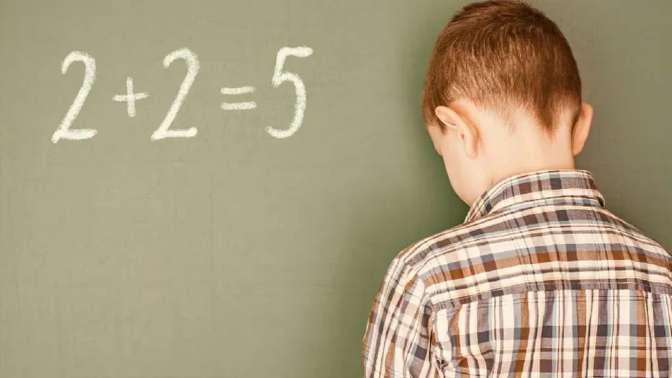 Un copil cu o cămașă în carouri stă cu spatele în fața unei table verzi pe care este scris 2+2=5 cu cretă albă.