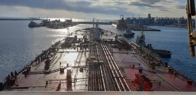 Privire de sus asupra punții unui vapor mare cu conducte metalice, în timp ce alte nave sunt vizibile pe apă în fundal.