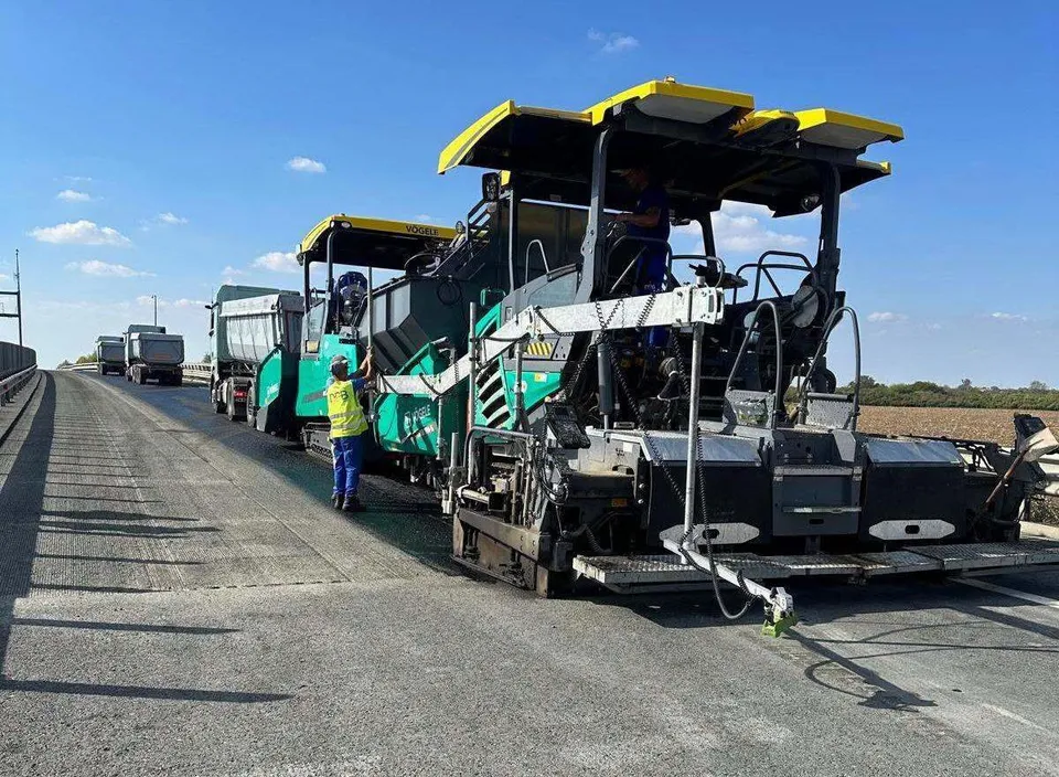 O mașină de asfaltat verde și galbenă stă pe o porțiune de drum asfaltat lângă un lucrător cu vestă reflectorizantă sub un cer senin.