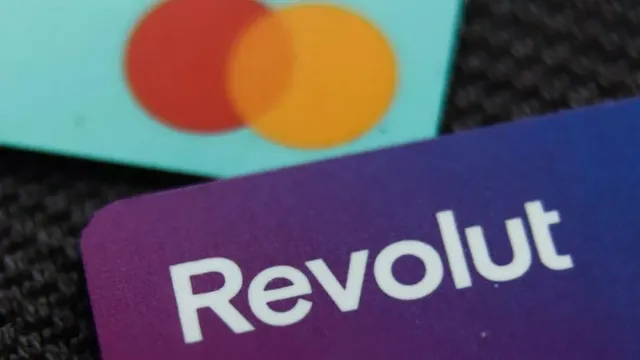 Un card bancar mov cu textul alb Revolut este plasat în prim-plan lângă un card cu două cercuri colorate pe un fundal albastru deschis.