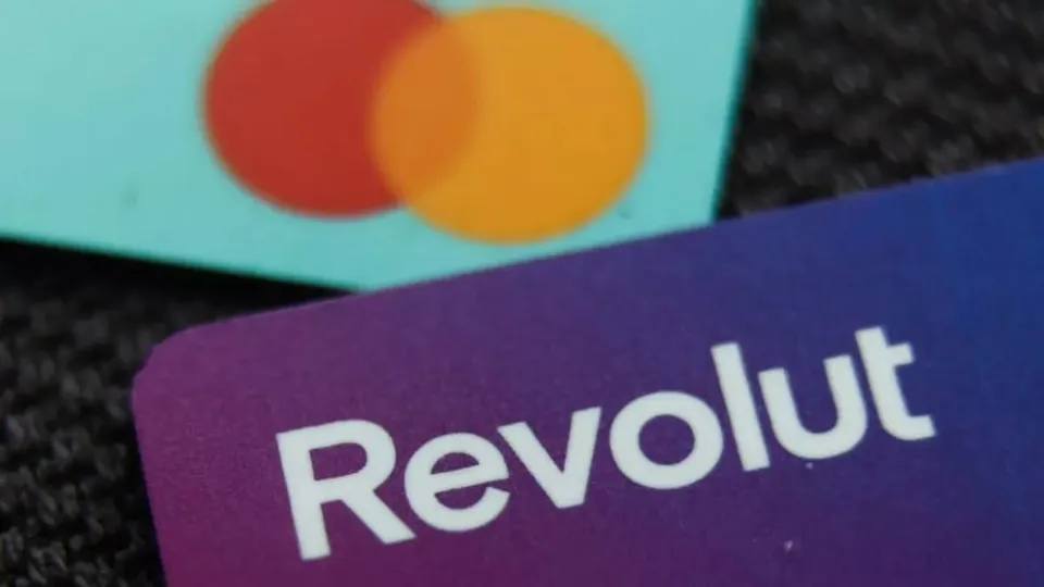 Un card bancar mov cu textul alb Revolut este plasat în prim-plan lângă un card cu două cercuri colorate pe un fundal albastru deschis.