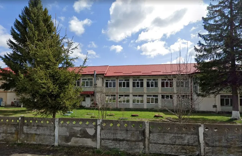 O clădire cu acoperiș roșu se află în spatele unui gard de beton și a unor copaci înalți sub un cer cu nori.