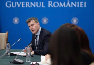 Un bărbat într-un costum închis la culoare stă la o masă cu microfoane și sticle de apă în fața unui fundal albastru scris.
