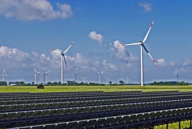 Rânduri de panouri solare sunt instalate pe un câmp verde sub un cer albastru cu nori, în prezența mai multor turbine eoliene albe.