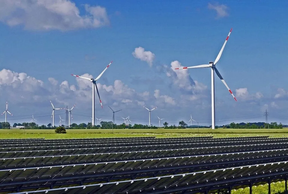 Rânduri de panouri solare sunt instalate pe un câmp verde sub un cer albastru cu nori, în prezența mai multor turbine eoliene albe.