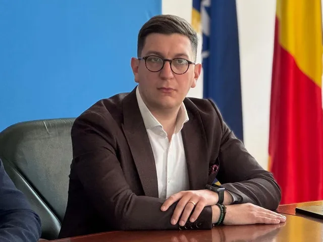 Un bărbat cu ochelari și sacou maro stă la o masă de lemn în fața unor steaguri.