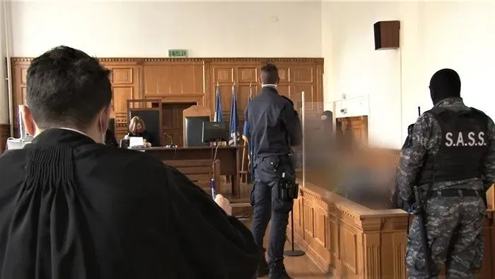 O persoană în robă neagră stă cu spatele, în timp ce doi bărbați în uniforme de poliție se află într-o sală de judecată cu mobilier din lemn.