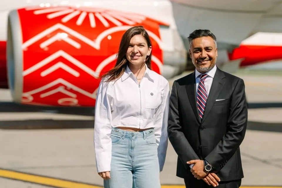 O femeie în cămașă albă și blugi deschiși stă lângă un bărbat în costum negru și cravată roz în fața unui motor de avion roșu.