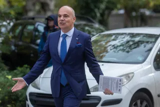 Un bărbat într-un costum albastru și cravată albastră merge pe lângă o mașină albă, ținând o broșură în mână și având mâna dreaptă întinsă.