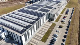 O vedere aeriană prezintă un clădire industrială lungă cu panouri solare pe acoperiș și un parc de parcare cu mașini aliniate pe o latură.