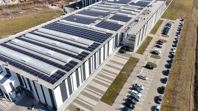 O vedere aeriană prezintă un clădire industrială lungă cu panouri solare pe acoperiș și un parc de parcare cu mașini aliniate pe o latură.