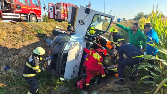 Mai mulți salvatori în uniforme colorate lucrează lângă o autoutilitară albă răsturnată într-un șanț, lângă un camion de pompieri roșu.