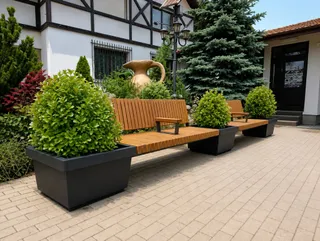 Bănci din lemn, jardiniere negre și arbuști verzi sunt așezate pe o curte pavată, lângă o clădire cu fațadă alb-negru.