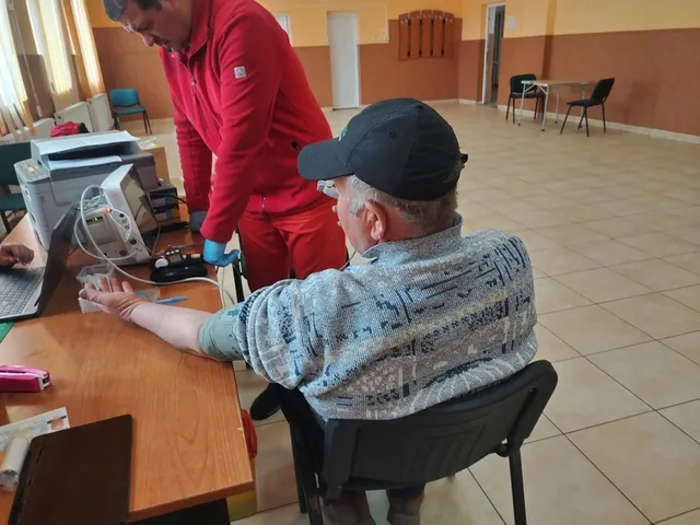 Un bărbat cu șapcă și cămașă modelată stă la o masă în timp ce un alt bărbat în haine roșii îi efectuează o procedură medicală.