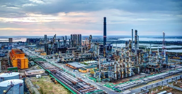 O vedere panoramică asupra unei rafinării industriale cu diverse coloane de distilare, conducte, depozite și un coș înalt sub un cer înnorat.
