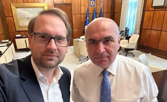 Un bărbat cu ochelari și sacou negru face un selfie alături de un bărbat cu cămașă albă și cravată albastră într-un birou.