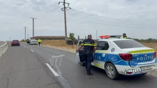 Un polițist stă lângă o mașină de poliție albă, albastră și galbenă pe un drum asfaltat, lângă alte vehicule parcate în depărtare.