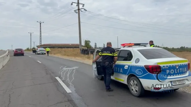 Un polițist stă lângă o mașină de poliție albă, albastră și galbenă pe un drum asfaltat, lângă alte vehicule parcate în depărtare.