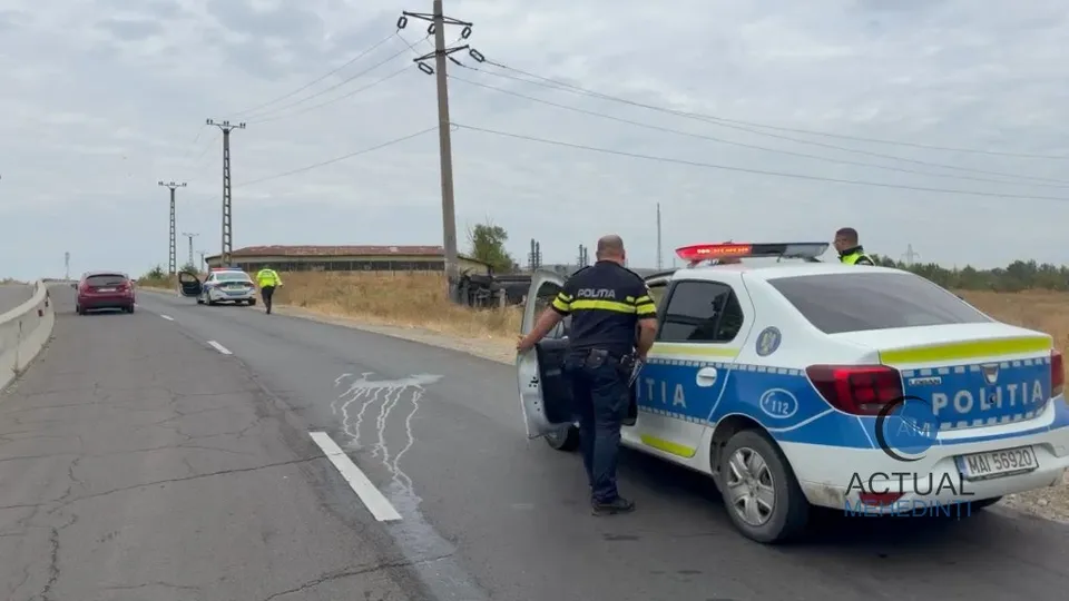 Un polițist stă lângă o mașină de poliție albă, albastră și galbenă pe un drum asfaltat, lângă alte vehicule parcate în depărtare.