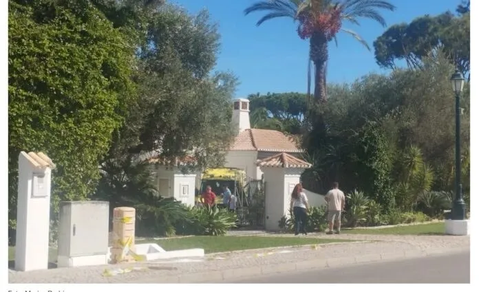 Oameni stau lângă un vehicul galben parcat pe un drum lângă o clădire cu acoperiș de țiglă și vegetație abundentă.