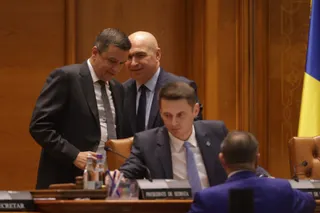 Trei bărbați în costume discută într-o sală de ședințe, cu un steag al Ucrainei vizibil în fundal. Unul dintre bărbați stă la masă, iar ceilalți doi stau în picioare.