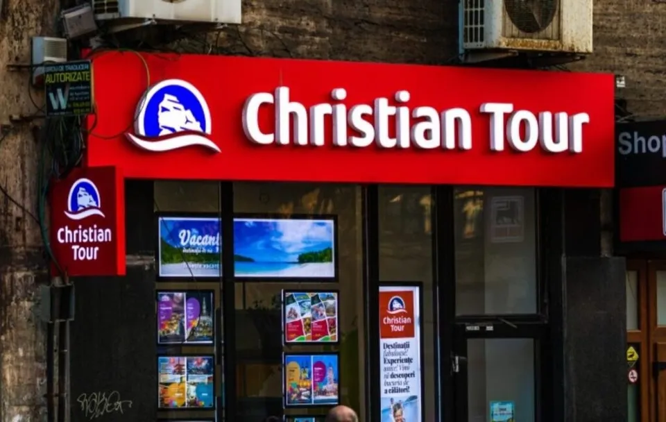 Un panou roșu cu textul alb Christian Tour și un logo albastru este montat deasupra unei vitrine de magazin cu afișe colorate.