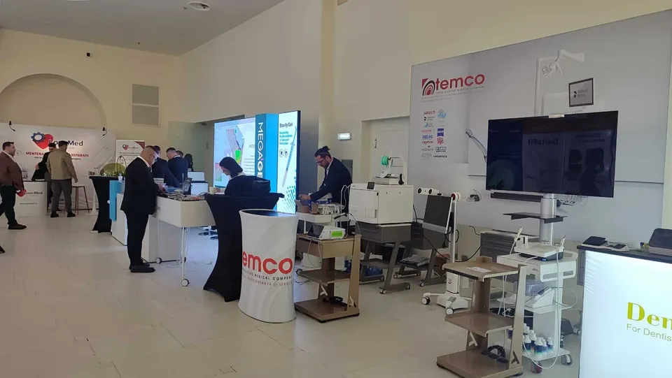 Oamenii stau în jurul unor standuri de expoziție cu echipamente medicale și ecrane într-o încăpere spațioasă cu pereți albi.