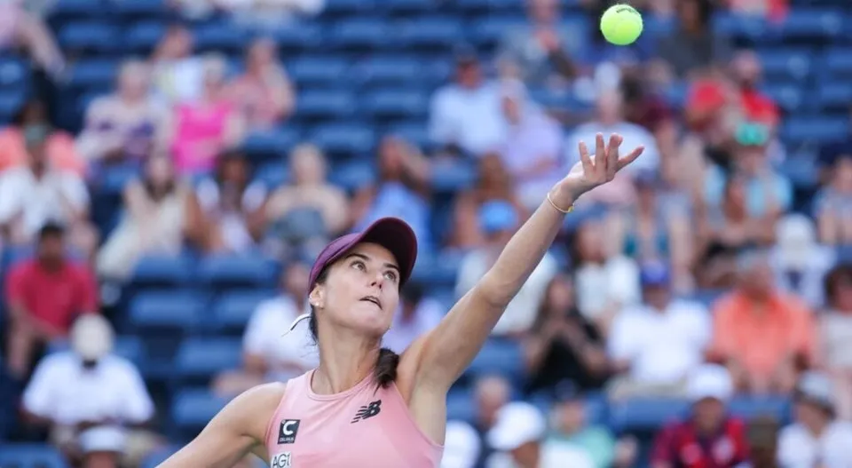 O femeie cu părul prins într-o coadă de cal ridică un braț spre o minge de tenis galbenă aflată în aer.