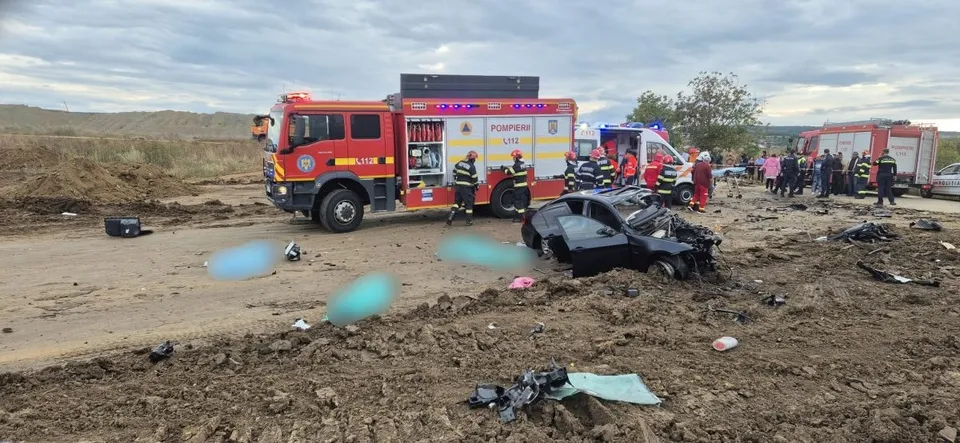 Un camion roșu, o ambulanță și un autoturism distrus stau pe un teren de culoare maro sub un cer înnorat.